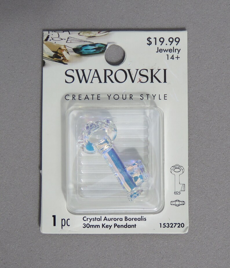 May include: A clear, iridescent crystal key pendant, 30mm in length, displayed on a white card with the Swarovski logo. The card reads "CREATE YOUR STYLE" and "Crystal Aurora Borealis".