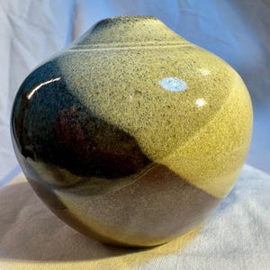 May include: A round ceramic vase with a glossy finish. The vase is decorated with three colors: a dark brown, a light brown, and a light yellow. The colors are arranged in a geometric pattern.