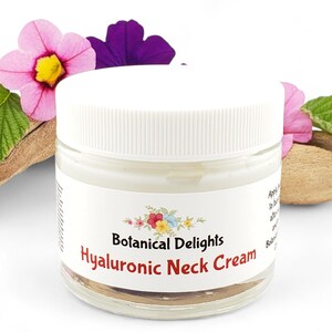 Op de afbeelding: Een witte pot met een wit deksel met Hyaluronzuur Halscr&egrave;me. De pot is gelabeld met "Botanical Delights Hyaluronic Neck Cream". De pot staat op een stuk drijfhout met roze en paarse bloemen.