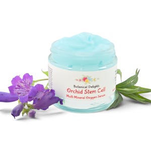 May include: A clear glass jar filled with light blue cream, featuring a white label that reads Botanical Delights Orchid Stem Cell Multi Mineral Oxygen Serum. The jar is positioned next to a cluster of vibrant purple flowers and green leaves.