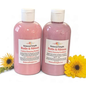 May include: Two 8 oz bottles of Botanical Delights Biotin & Hibiscus hair care products, one shampoo and one conditioner, featuring pink liquid, white caps, and labels with floral accents, accompanied by two yellow daisy-like flowers.