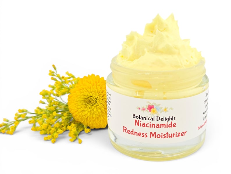 May include: A clear glass jar filled with a yellow cream, labeled "Botanical Delights Niacinamide Redness Moisturizer." A yellow flower and sprigs are next to the jar. The background is white.