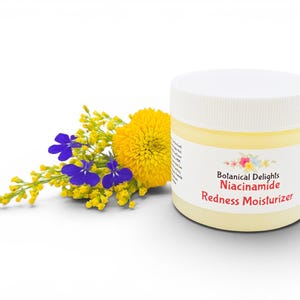 May include: A jar of Botanical Delights Niacinamide Redness Moisturizer with a white lid and a yellow label. The jar is next to a bouquet of yellow and purple flowers. The background is white.