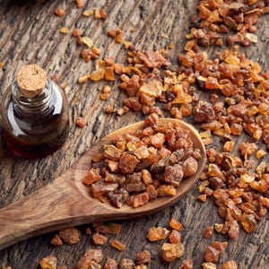 May include: A small glass bottle with a cork stopper sits beside a wooden spoon filled with amber-coloured resin. The resin and bottle are on a textured wooden surface, with scattered pieces of resin.