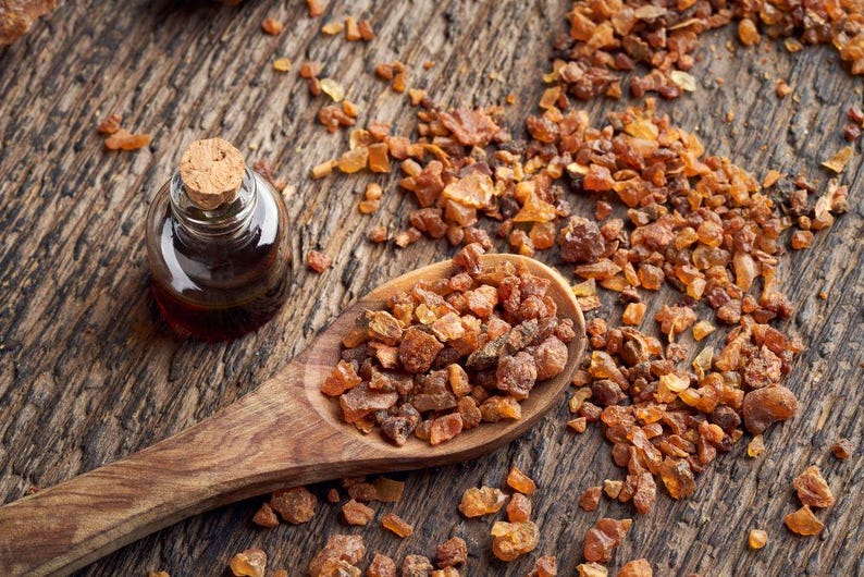May include: A small glass bottle with a cork stopper sits beside a wooden spoon filled with amber-coloured resin. The resin and bottle are on a textured wooden surface, with scattered pieces of resin.