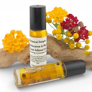 May include: Two glass roll-on bottles of Botanical Delights Frankincense & Myrrh Resin Infused Oil with black caps. The bottles are filled with a golden liquid. The bottles are next to a piece of driftwood and a bouquet of yellow and red flowers.