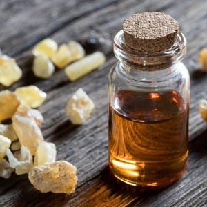 May include: A small glass bottle with a cork stopper filled with amber liquid, alongside several pieces of amber-coloured resin. The items are on a weathered wooden surface, suggesting a natural product.