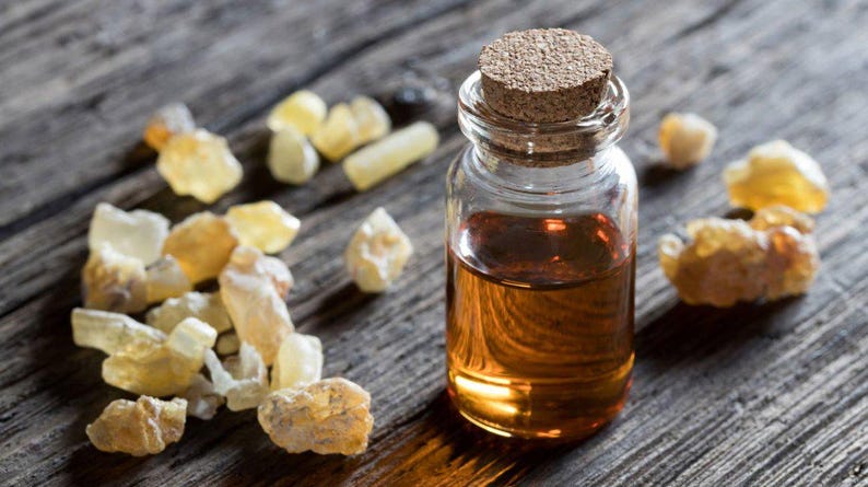 May include: A small glass bottle with a cork stopper filled with amber liquid, alongside several pieces of amber-coloured resin. The items are on a weathered wooden surface, suggesting a natural product.