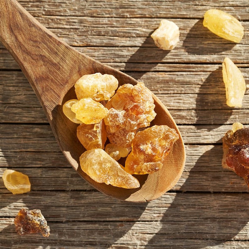May include: A wooden spoon holding various pieces of amber-coloured resin. The resin pieces are of different sizes and shapes, some appearing translucent. The spoon sits on a weathered wooden surface, with more resin pieces scattered around.