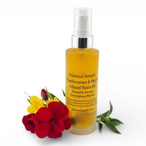May include: A bottle of yellow resin oil with a silver pump, labelled "Botanical Delights Frankincense & Myrrh Infused Resin Oil." Red and yellow flowers and green leaves are arranged near the bottle. The bottle is made of clear glass.