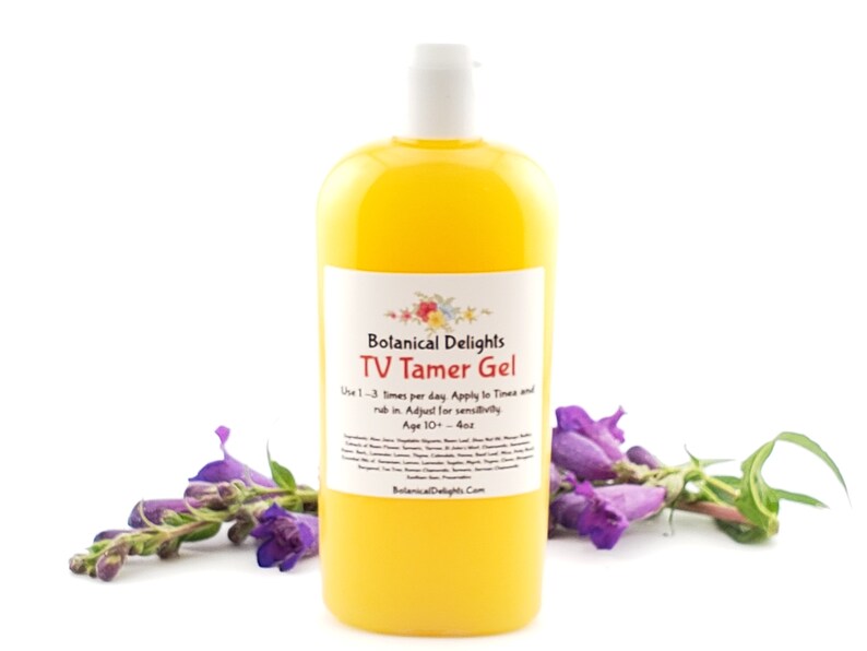 May include: A yellow bottle of TV Tamer Gel by Botanical Delights. The bottle is labeled with the product name and instructions for use. The bottle is sitting on a white surface with purple flowers in the foreground.
