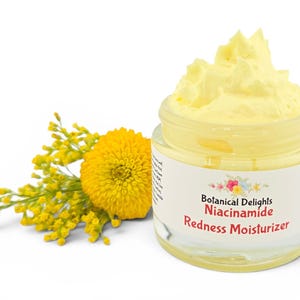 May include: A clear glass jar of yellow moisturizer with the text "Botanical Delights Niacinamide Redness Moisturizer." A yellow flower and sprig of yellow flowers are to the left of the jar. The background is white.