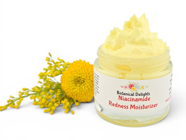 May include: A clear glass jar of yellow moisturizer with the text "Botanical Delights Niacinamide Redness Moisturizer." A yellow flower and sprig of yellow flowers are to the left of the jar. The background is white.