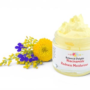 May include: A jar of yellow moisturizer with the text "Botanical Delights Niacinamide Redness Moisturizer." A yellow and blue floral arrangement is to the left of the jar. The jar is clear with a white label.