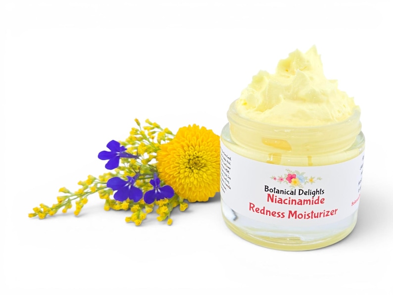 May include: A jar of yellow moisturizer with the text "Botanical Delights Niacinamide Redness Moisturizer." A yellow and blue floral arrangement is to the left of the jar. The jar is clear with a white label.