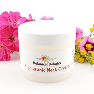 Op de afbeelding: Een witte pot met een wit deksel en een etiket met de tekst "Botanical Delights Hyaluronic Neck Cream". De pot is omringd door roze en gele bloemen.