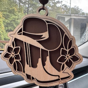 May include: A wooden car air freshener with a cowboy boot and hat design, accented with two floral elements. The air freshener is suspended by a string of wooden beads. The word "Nana" is written on the bottom right.