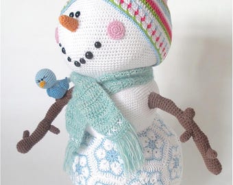 Könnte beinhalten: Ein gehäkelter Schneemann mit einem blauen Vogel, der auf einem Ast sitzt. Der Schneemann trägt einen bunten gestreiften Hut, einen hellblauen Schal und ein weiß-blaues Schneeflockenmuster auf seinem Körper.