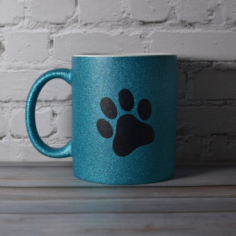 Puede incluir: Una taza de color azul cerceta brillante con un acabado de purpurina y un gr&aacute;fico grande de una huella negra centrado en la parte frontal. La taza cuenta con un asa curva en el lado izquierdo y un borde interior blanco y liso.