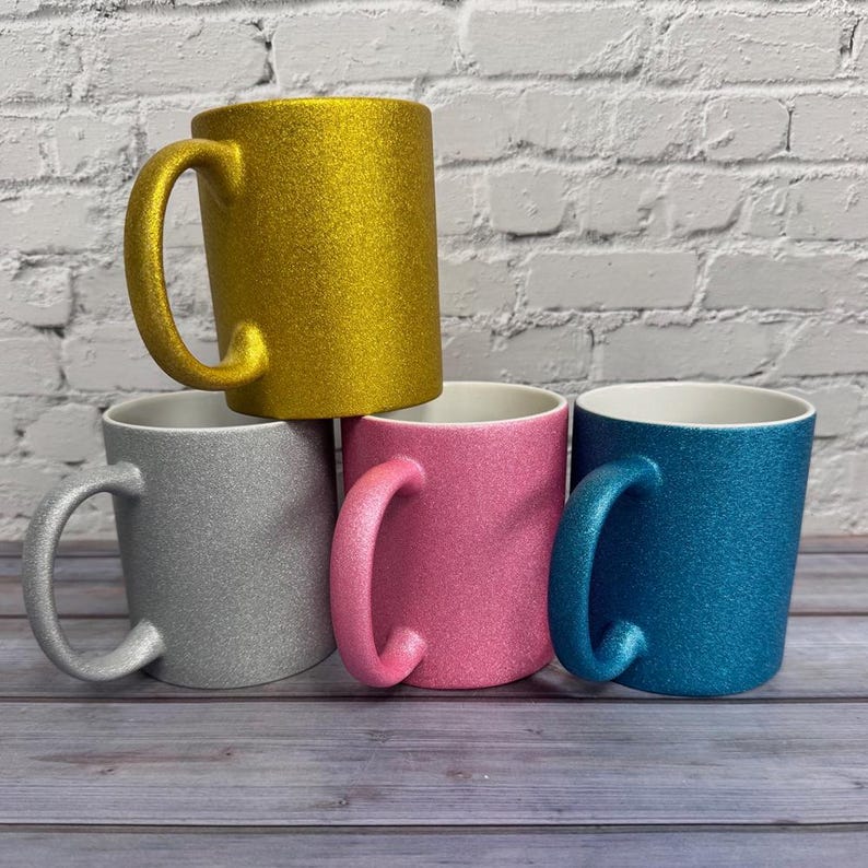 Puede incluir: Un conjunto de cuatro tazas de cer&aacute;mica con acabado brillante. El set incluye una taza dorada colocada sobre otras tres de color plateado, rosa y azul. Cada taza tiene un asa curva y una superficie interior blanca y lisa.