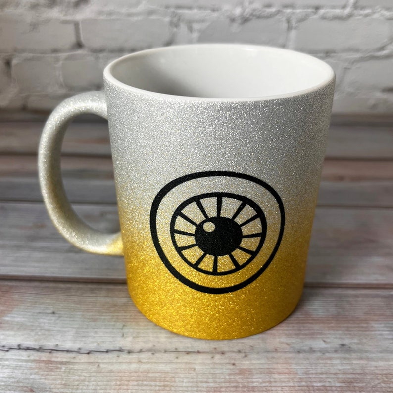 Puede incluir: Una taza de cer&aacute;mica con un acabado degradado brillante que pasa de plateado en la parte superior a dorado en la inferior. El centro de la taza muestra un gr&aacute;fico negro llamativo de un ojo estilizado con una pupila circular y l&iacute;neas de iris.