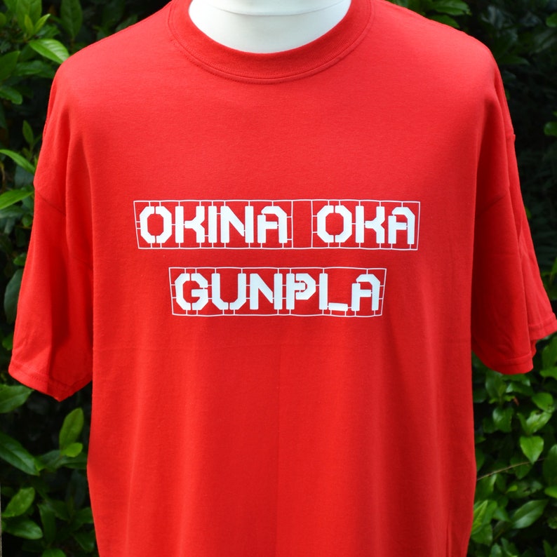 May include: Red t-shirt with white text that reads "OKINA OKA GUNPLA" in a blocky font.