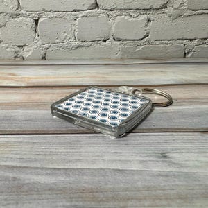 May include: A clear plastic keychain with a square-shaped charm featuring a white background with a repeating pattern of blue circles. The keychain has a silver metal ring.