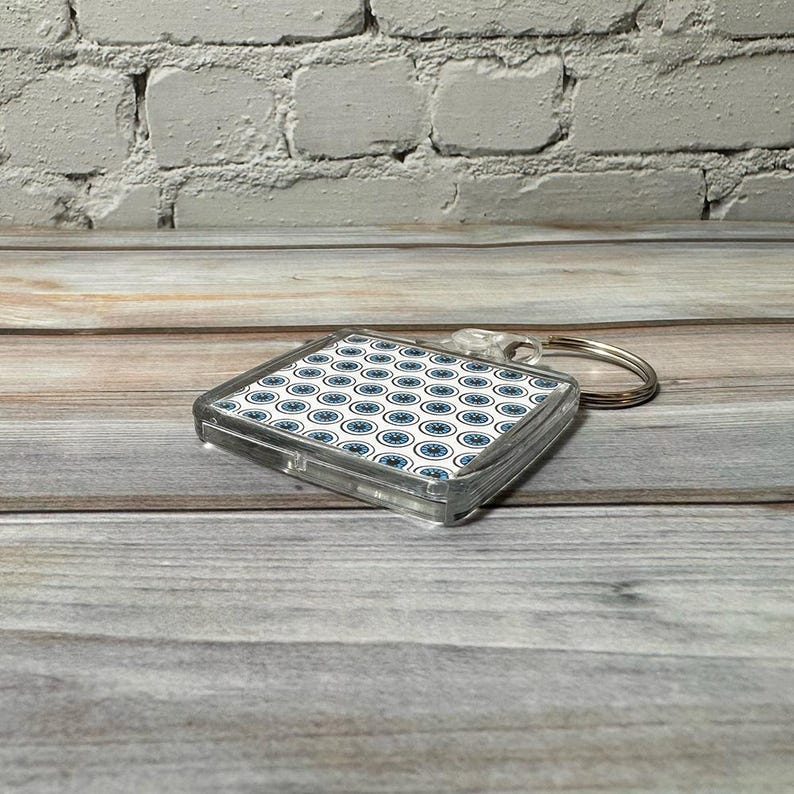 May include: A clear plastic keychain with a square-shaped charm featuring a white background with a repeating pattern of blue circles. The keychain has a silver metal ring.