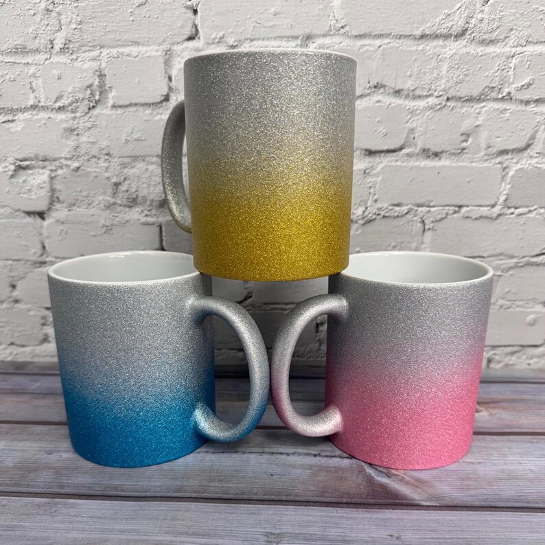 Puede incluir: Tres tazas de cer&aacute;mica con acabado de purpurina. La taza superior presenta un degradado de plata a oro. La taza inferior izquierda muestra un degradado de plata a azul, mientras que la inferior derecha muestra un degradado de plata a rosa.