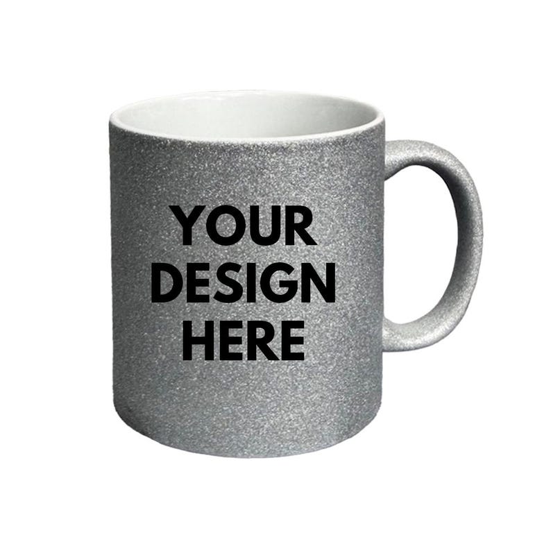 Puede incluir: Una taza de cer&aacute;mica con acabado de purpurina plateada, interior blanco y asa en el lado derecho. La parte frontal de la taza presenta el texto negro "YOUR DESIGN HERE" impreso en una fuente sans-serif negrita, centrado en la superficie.