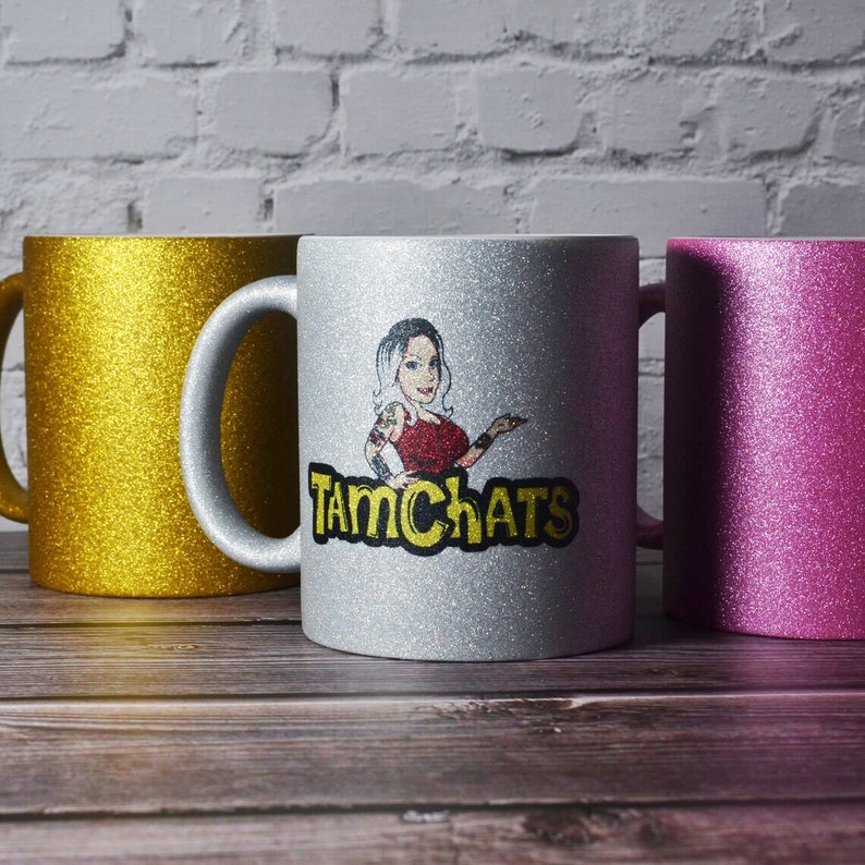 Puede incluir: Tres tazas de cer&aacute;mica con purpurina en oro, plata y rosa. La taza plateada central presenta un gr&aacute;fico de una persona con cabello oscuro y tatuajes con una blusa roja, sobre el texto amarillo y negro que dice TAMCHATS.