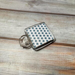 May include: A clear plastic keychain with a square-shaped charm featuring a white background with a repeating pattern of blue eyes.
