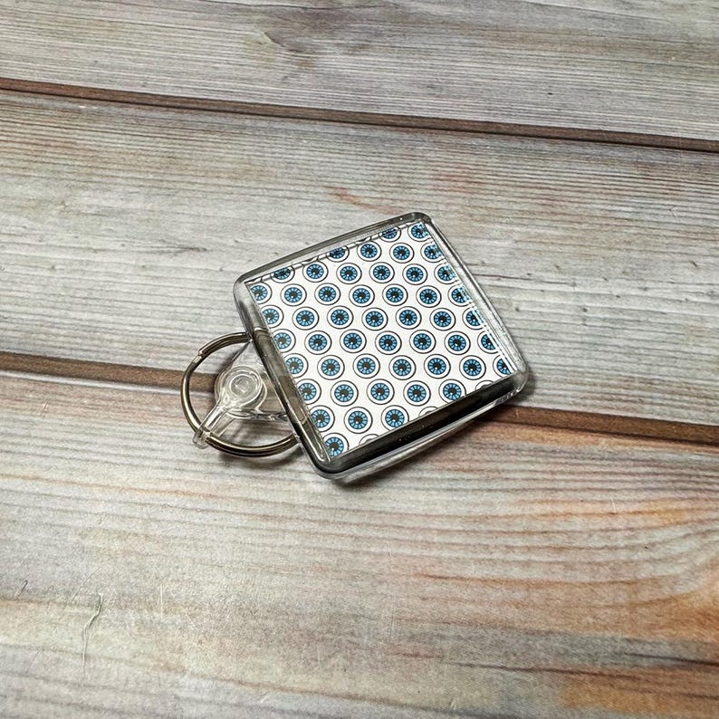 May include: A clear plastic keychain with a square-shaped charm featuring a white background with a repeating pattern of blue eyes.