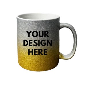Puede incluir: Una taza de cer&aacute;mica con un acabado degradado brillante que pasa de plateado en la parte superior a dorado en la inferior. El texto "YOUR DESIGN HERE" est&aacute; impreso en letras negras negritas sin remates en el centro frontal de la taza.