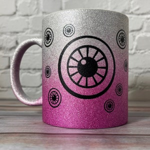 Puede incluir: Una taza con acabado brillante y degradado de color plata a rosa. La superficie est&aacute; decorada con m&uacute;ltiples gr&aacute;ficos circulares negros que parecen ojos de distintos tama&ntilde;os, incluyendo un dise&ntilde;o central grande y varios motivos menores.