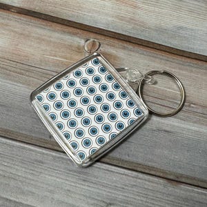 May include: A clear plastic keychain with a square-shaped charm featuring a repeating pattern of blue eyes on a white background.