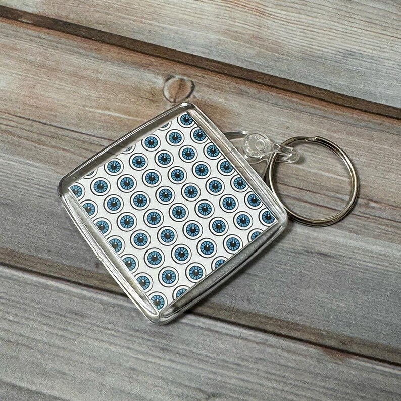 May include: A clear plastic keychain with a square-shaped charm featuring a repeating pattern of blue eyes on a white background.