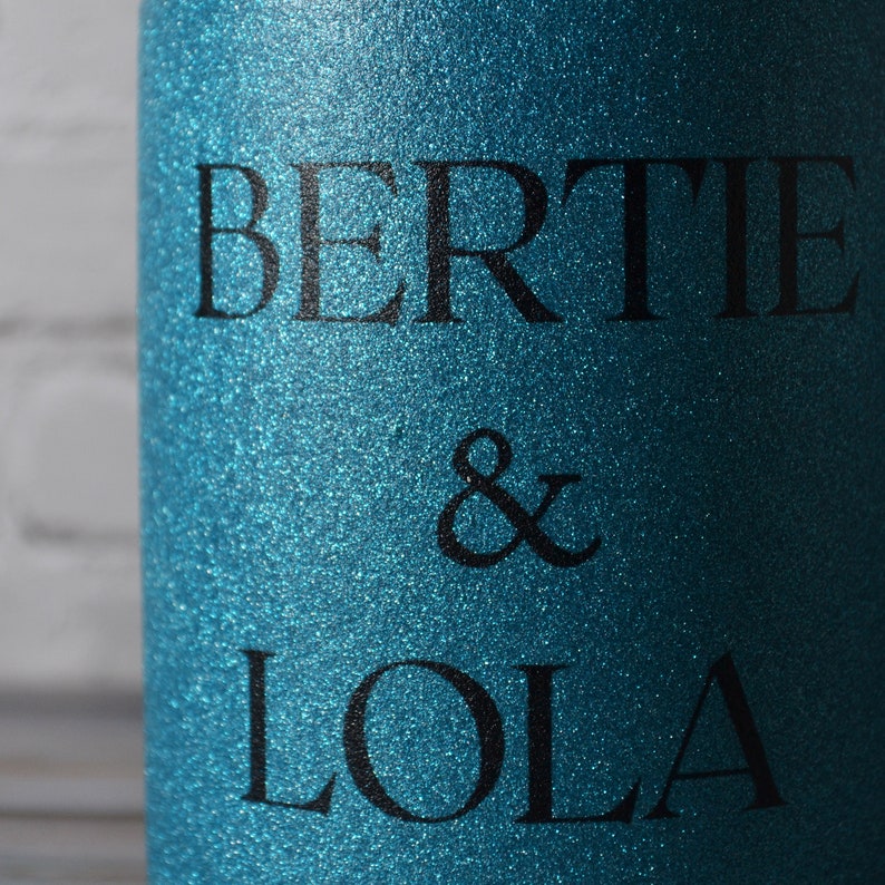 Puede incluir: Un objeto cil&iacute;ndrico con un acabado de purpurina en color verde azulado vibrante. La superficie presenta el texto negro "BERTIE & LOLA" impreso en una fuente negrita sin remates. La textura de purpurina es uniforme en toda la superficie.