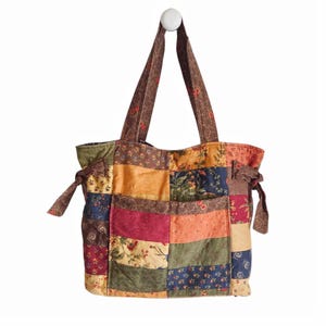 May include: A patchwork tote bag constructed from various fabric squares featuring floral and solid patterns in shades of burgundy, navy blue, olive green, mustard yellow, and orange. The bag includes two shoulder straps and side tie details.