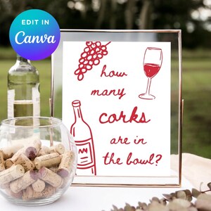 May include: A framed sign with red illustrations of grapes, a wine glass, and a bottle, along with the text "how many corks are in the bowl?" A glass bowl filled with corks sits in front of the sign. A bottle of wine is in the background.