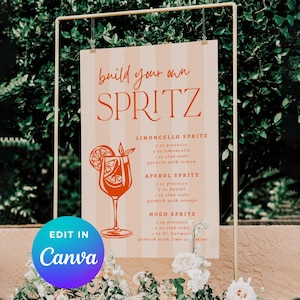 May include: A sign with the words "build your own SPRITZ" in orange script. The sign includes drink recipes for Limoncello, Aperol, and Hugo Spritz. The sign is displayed on a gold metal frame, with a Canva logo in the bottom left corner.