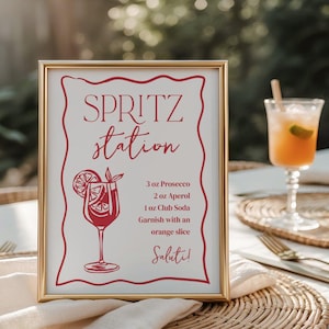 May include: A framed sign with a gold border displays a Spritz Station recipe. The sign includes the text "SPRITZ station" in a decorative font, along with the ingredients: 3 oz Prosecco, 2 oz Aperol, and 1 oz Club Soda. The sign is garnished with an orange slice and the word "Saluti!"