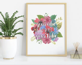 May include: A framed print with the text "your vibe attracts your tribe" in white script, surrounded by a watercolor floral arrangement in shades of pink, red, blue, and green. The frame is gold.