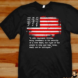 Può includere: T-shirt nera con un design a bandiera americana invecchiata con strisce rosse e bianche e sagome di pistole bianche. La maglietta include il testo del Secondo Emendamento: "Una milizia ben regolamentata...non sarà violata."