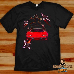 May include: A black short-sleeve t-shirt featuring a graphic print of a red sports car with a ninja standing on its roof. The ninja holds a sword, and several metallic throwing stars are depicted flying around the car with motion trails.