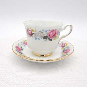 Könnte beinhalten: Weißes Tee-Service mit einer Tasse und Untertasse, verziert mit rosa Rosen, gelben und blauen Blumen und goldenen Rändern. Die Tasse hat einen gebogenen Henkel und steht auf einer passenden Untertasse.