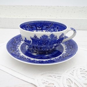May include: A white ceramic teacup and matching saucer featuring a detailed blue willow pattern. The design depicts traditional pagodas, trees, and bridges in a classic transferware style. The cup has a curved handle and a wide rim.