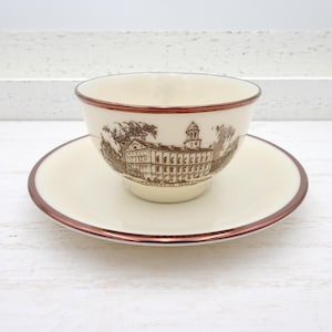 Puede incluir: Juego de taza y platillo de té color crema con una ilustración de edificio marrón. La taza y el platillo tienen un borde marrón. La taza de té está sobre el platillo. El juego está sobre una superficie blanca.