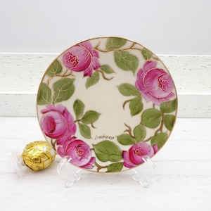 May include: A decorative plate featuring hand-painted pink roses with green leaves and gold accents. The plate has a gold rim and is displayed on a clear stand. The artist's signature, "J. Sechner", is visible on the plate.