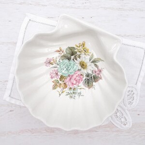 Puede incluir: Un plato de cerámica blanca con forma de concha marina, que presenta un arreglo floral central con flores rosas, verde menta, amarillas y blancas rodeadas de hojas verdes. El plato tiene un borde festoneado y un acabado brillante sobre tela.